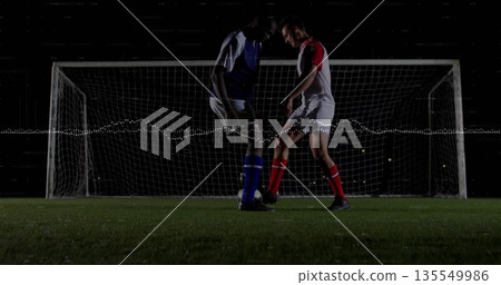 Two soccer players in blue and red kits chasing ball under stadium lights, with goal net 135549986