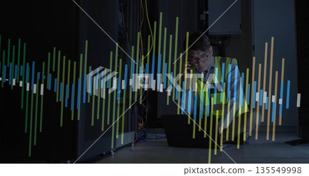 Technician kneeling among data center racks wearing safety vest and using smartphone and laptop 135549998