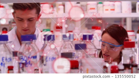Examining researchers wearing white coats analyzing sample on bench in lab, with reagent bottles Examining researchers wearing white coats analyzing sample on bench in lab, with reagent bottles 135550052