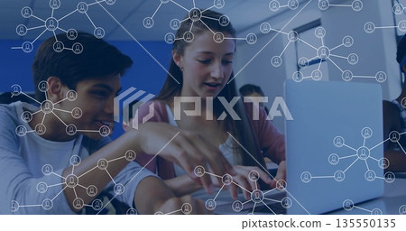 Typing teenage girl and pointing bi-cultural boy at classroom desk, showing laptop network icons Typing teenage girl and pointing bi-cultural boy at classroom desk, showing laptop network icons 135550135