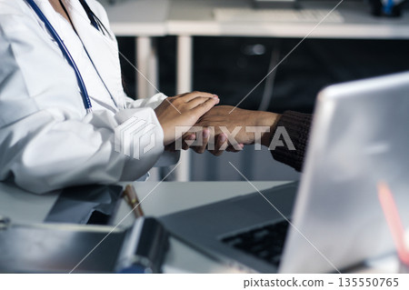 Senior patient lying in hospital bed receiving comforting touch from young female nurse, hands gently clasped together, medical care setting visible Senior patient lying in hospital bed receiving comforting touch from young female nurse, hands gently clasped together, medical care setting visible 135550765