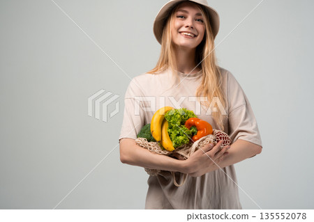 Happy young woman holding fresh vegetables and fruit in reusable mesh bag, plastic free food shopping and sustainability 135552078