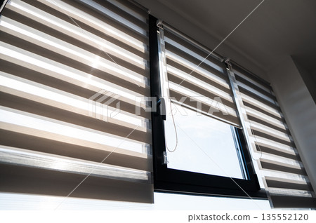 Modern apartment window with zebra roller blinds and chain control system creating balanced natural light indoors Modern apartment window with zebra roller blinds and chain control system creating balanced natural light indoors 135552120