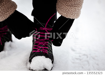 A close-up shot of a person's hands tying pink laces on black hiking boots in the snow. Authentic image captures the preparation for winter adventure. A close-up shot of a person's hands tying pink laces on black hiking boots in the snow. Authentic image captures the preparation for winter adventure. 135552323