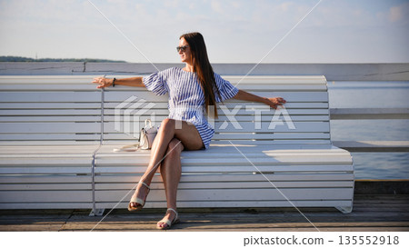 Young woman sitting on white bench by the sea on sunny day Young woman sitting on white bench by the sea on sunny day 135552918