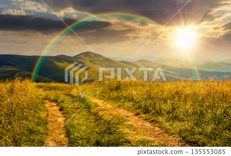 dirt road through grassy alpine meadow on the ridge at sunset. beautiful summer landscape in green mountains under cloudy sky in evening light. timeless nostalgia book cover with rainbow dirt road through grassy alpine meadow on the ridge at sunset. beautiful summer landscape in green mountains under cloudy sky in evening light. timeless nostalgia book cover with rainbow 135553085