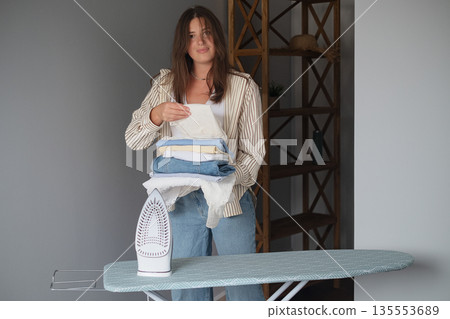 Cozy home scene with girl ironing fresh clothes with a steam iron and neatly stacking them creating a clean organized wardrobe Cozy home scene with girl ironing fresh clothes with a steam iron and neatly stacking them creating a clean organized wardrobe 135553689