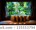 Window corner decorated with square pillows on sofa with blurry background of tropical garden. 135553794