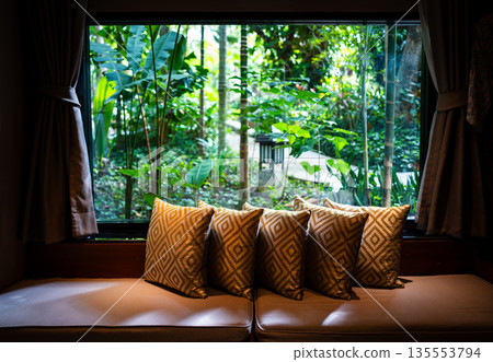 Window corner decorated with square pillows on sofa with blurry background of tropical garden. Window corner decorated with square pillows on sofa with blurry background of tropical garden. 135553794