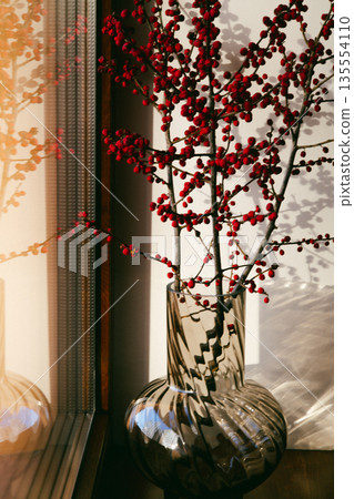 Minimalist interior still life featuring a transparent glass vase with red berry branches placed on a windowsill. A play of shadows and light on wall. 135554110