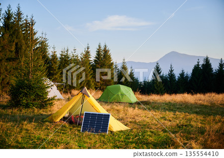 Solar panel in grassy field with tents in background highlights eco-friendly camping setup, surrounded by dense evergreen trees in evening. Integration of renewable energy in outdoor activities. Solar panel in grassy field with tents in background highlights eco-friendly camping setup, surrounded by dense evergreen trees in evening. Integration of renewable energy in outdoor activities. 135554410