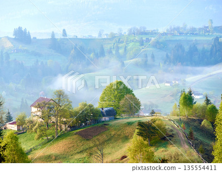 Serene countryside morning with mist rolling over gentle hills. Sunlight filters through, highlighting cozy farmhouse surrounded by lush trees. Scattered homes and winding paths, mist-covered valleys. Serene countryside morning with mist rolling over gentle hills. Sunlight filters through, highlighting cozy farmhouse surrounded by lush trees. Scattered homes and winding paths, mist-covered valleys. 135554411