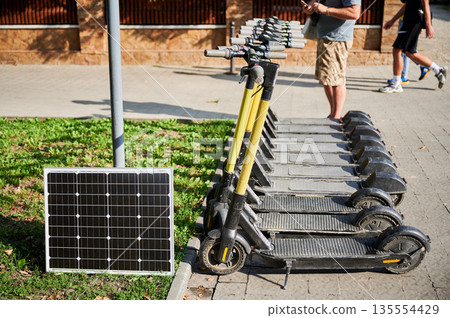 Row of electric scooters parked beside solar panel in outdoor urban area. Integration of solar power as sustainable energy source for charging electric scooters, promoting eco-friendly transportation. Row of electric scooters parked beside solar panel in outdoor urban area. Integration of solar power as sustainable energy source for charging electric scooters, promoting eco-friendly transportation. 135554429