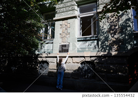 Woman holding photovoltaic solar panel in front of historical building, wearing plaid shirt and sunglasses. Concept of integration of sustainable renewable energy sources into old architecture. 135554432