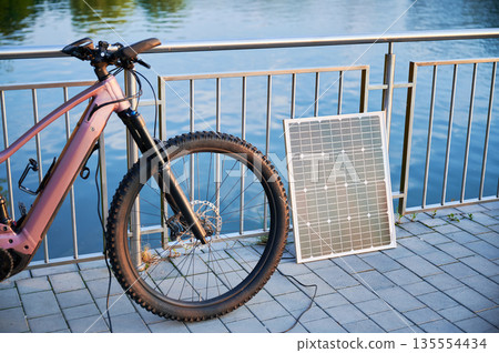 Pink electric mountain bike parked beside lakeside railing, connected to solar panel for charging. Bicycle's sleek design and rugged tires highlighted, with tranquil lake and golden light. 135554434