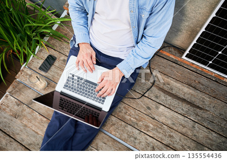 Top view of man works on laptop on lap and smartphone, sits on wooden deck. Solar panel nearby, emphasizing sustainable, modern eco-friendly workspace that combines technology and sustainability. Top view of man works on laptop on lap and smartphone, sits on wooden deck. Solar panel nearby, emphasizing sustainable, modern eco-friendly workspace that combines technology and sustainability. 135554436