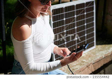 Woman connecting smartphone to photovoltaic solar panel. Cropped view. Integration of sustainable renewable energy into everyday life, demonstrating practical use of solar power for charging devices. Woman connecting smartphone to photovoltaic solar panel. Cropped view. Integration of sustainable renewable energy into everyday life, demonstrating practical use of solar power for charging devices. 135554442