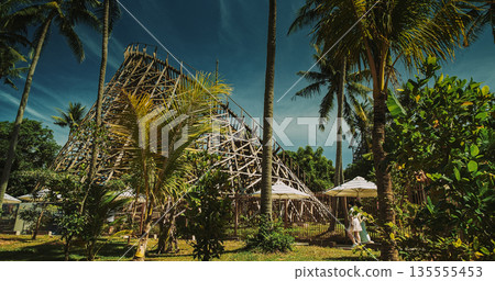 Scenic wooden boardwalk path beneath massive rollercoaster in tropical forest. Scenic wooden boardwalk path beneath massive rollercoaster in tropical forest. 135555453