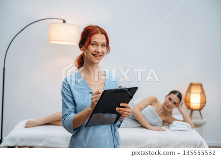 Holding notepad. Female masseur near woman lying down in spa salon Holding notepad. Female masseur near woman lying down in spa salon 135555532
