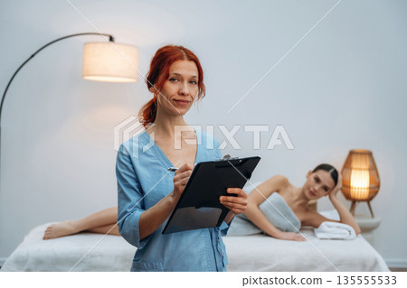 Holding notepad. Female masseur near woman lying down in spa salon Holding notepad. Female masseur near woman lying down in spa salon 135555533