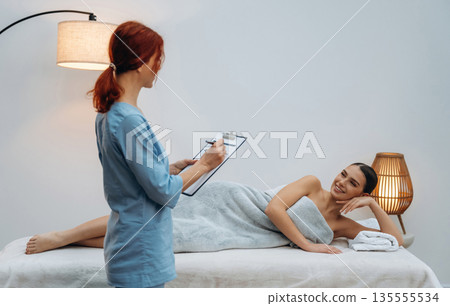Writing in the notepad. Female masseur near woman lying down in spa salon Writing in the notepad. Female masseur near woman lying down in spa salon 135555534