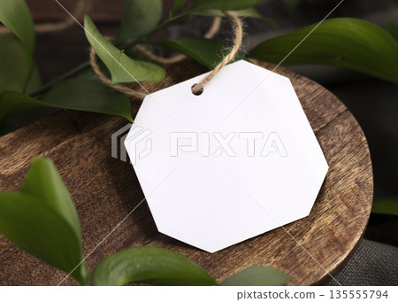 Blank gift tag mockup on brown wooden tray near green leaves and grey fabric closeup Blank gift tag mockup on brown wooden tray near green leaves and grey fabric closeup 135555794