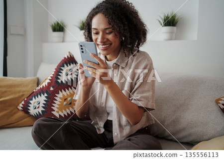 Woman With Bright Smile Reviews Playful Message On Her Smartphone Surrounded By Cozy Decor And Cushions 135556113