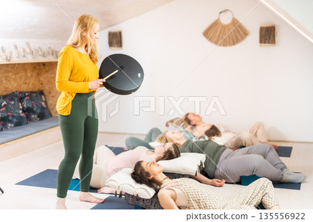 Group therapy session with Tibetan drum for group of women lying on wooden floor surrounded by copper bowls. Meditation and relaxation 135556922
