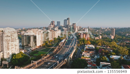 Australia, Sydney: Sydney's skyline at sunrise, skyscrapers, cars driving highway road, residential buildings, and green spaces. Aerial drone flight over modern cityscape, urban architecture Australia, Sydney: Sydney's skyline at sunrise, skyscrapers, cars driving highway road, residential buildings, and green spaces. Aerial drone flight over modern cityscape, urban architecture 135556949