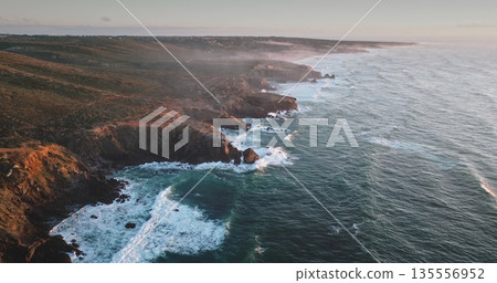 Portugal, Lisbon: Dramatic rugged cliffs and verdant hills lining the Atlantic Ocean coastline, with golden hour sunlight illuminating the landscape, waves crash against the shore. Aerial drone flight Portugal, Lisbon: Dramatic rugged cliffs and verdant hills lining the Atlantic Ocean coastline, with golden hour sunlight illuminating the landscape, waves crash against the shore. Aerial drone flight 135556952