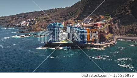 Tenerife: Modern residential buildings and resorts on rugged coastline of Atlantic Ocean, waves breaking on rocky shore and winding road ascending the volcanic mountain landscape. Aerial view panorama Tenerife: Modern residential buildings and resorts on rugged coastline of Atlantic Ocean, waves breaking on rocky shore and winding road ascending the volcanic mountain landscape. Aerial view panorama 135556959