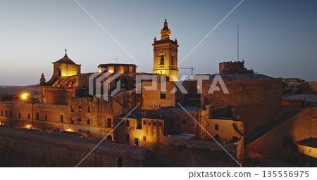 Malta, Comino island: Warm night lights illuminating the Cittadella in Gozo, Malta, highlight the medieval architecture of the fortified city at dusk. Aerial drone flight panorama 135556975