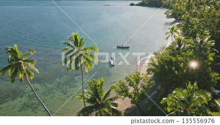 Two boats resting on the shallow turquoise water close to a pristine sandy beach lined with lush green palm trees and dense jungle on a sunny day in Fiji. 135556976