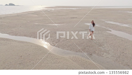 Woman enjoying leisurely barefoot walk on vast sandy beach at low tide, explore receding waters and textured patterns under the warm sunlight during a relaxing ocean vacation. Aerial drone flight 135556982