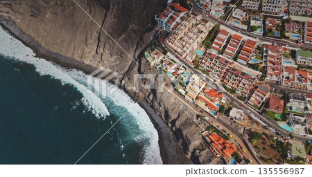 Tenerife: Los Gigantes dramatic rocky coastline town, Canary Islands, dark blue ocean waves breaking on the volcanic mountain hill shore. Resort hotel residential buildings top down, aerial flight 135556987