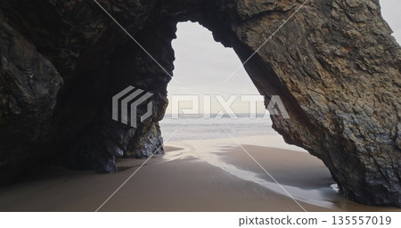 Portugal, Lisbon: rock arch at Praia da Adraga beach framing the Atlantic ocean, waves breaking on wet sand shore under cloudy sky. Low angle drone flight in mountain cliff hole. Nature background 135557019