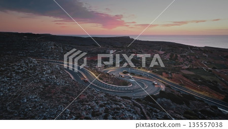 Malta: Breathtaking aerial view of cars driving along winding coastal road in Malta during a vibrant sunset, showcasing the island's beautiful landscape and the Mediterranean Sea. Drone flight Malta: Breathtaking aerial view of cars driving along winding coastal road in Malta during a vibrant sunset, showcasing the island's beautiful landscape and the Mediterranean Sea. Drone flight 135557038