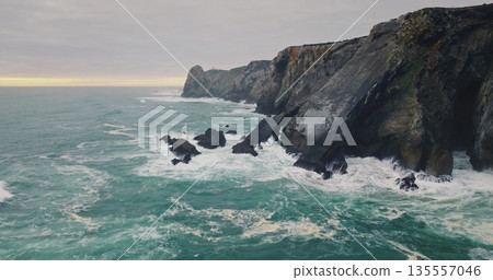 Portugal, Lisbon: dramatic Praia da Adraga beach coast showing powerful ocean waves crashing against dark, rugged cliffs and rocky formations, creating a moody coastal seascape. Aerial drone flight Portugal, Lisbon: dramatic Praia da Adraga beach coast showing powerful ocean waves crashing against dark, rugged cliffs and rocky formations, creating a moody coastal seascape. Aerial drone flight 135557046