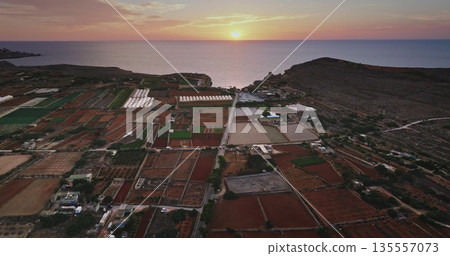Malta: Aerial view of colorful fields and greenhouses covering the land in Malta with the Mediterranean Sea in the background at sunset. Drone flight 135557073