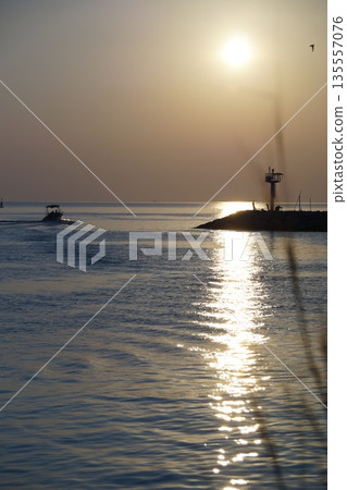 The golden sun rises from behind the blue sky on a stone pier and lighthouse, against a backdrop of trees and grass.Dawn of the golden sun. The golden sun rises from behind the blue sky on a stone pier and lighthouse, against a backdrop of trees and grass.Dawn of the golden sun. 135557076