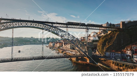Portugal: Dom Luis I Bridge spans Douro River in Porto, with commuter train on upper deck and traffic below, framed by historic riverfront buildings at golden sunset hour. Aerial view drone 135557079