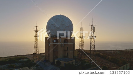 Aerial military ground radio station with radar antenna shelters and radio towers at sunset. The spherical radar dome stands prominently against the serene coastal backdrop. Drone flight 135557103