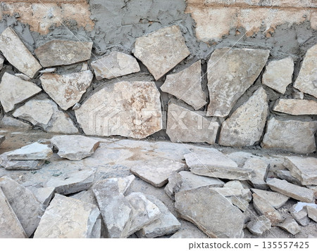 Workers are placing natural stone pieces on a wall to finish a construction project. Some stone pieces are on the ground, ready for use. The work is taking place outside. Workers are placing natural stone pieces on a wall to finish a construction project. Some stone pieces are on the ground, ready for use. The work is taking place outside. 135557425