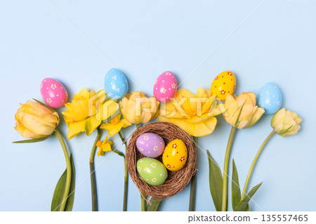 Easter and spring concept. Easter colored painted eggs in bird nest with yellow daffodils, white tulips on blue background with space for text and greetings. Spring background Easter and spring concept. Easter colored painted eggs in bird nest with yellow daffodils, white tulips on blue background with space for text and greetings. Spring background 135557465