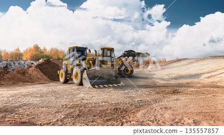 Two loaders work together on a construction site, moving soil and materials outdoors under a blue, partly cloudy sky Two loaders work together on a construction site, moving soil and materials outdoors under a blue, partly cloudy sky 135557857