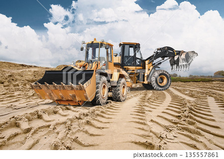 Heavy equipment two loaders is conducting construction work, planning the future construction site. A large pile of sand is visible in the background under a cloudy sky 135557861