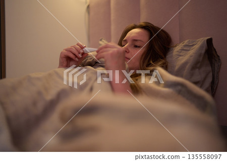 Female lying in bed examining thermometer indoors Female lying in bed examining thermometer indoors 135558097