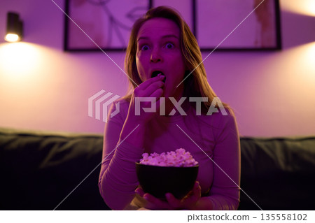Female eating popcorn on sofa illuminated by colored room lights Female eating popcorn on sofa illuminated by colored room lights 135558102