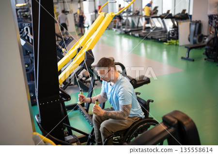 Motivated disabled man in wheelchair working on upper body strength with vertical fitness machine. Motivated disabled man in wheelchair working on upper body strength with vertical fitness machine. 135558614
