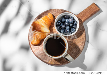 Freshly brewed espresso coffee in a cup, a croissant and blueberries on a wooden board Freshly brewed espresso coffee in a cup, a croissant and blueberries on a wooden board 135558943
